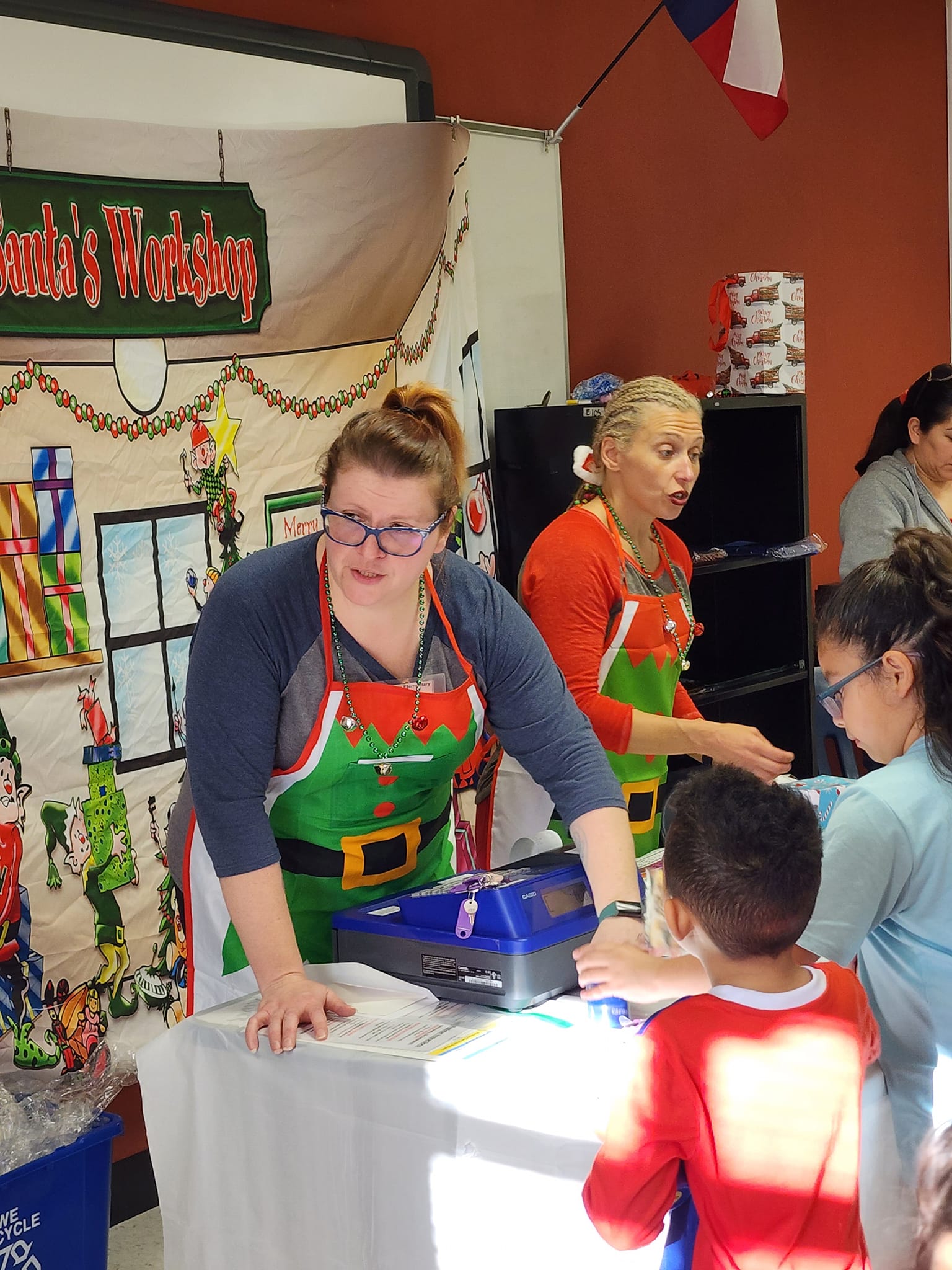 Two female PTA members dressed in holiday apron's helping children at the Elf Shelf store. 
