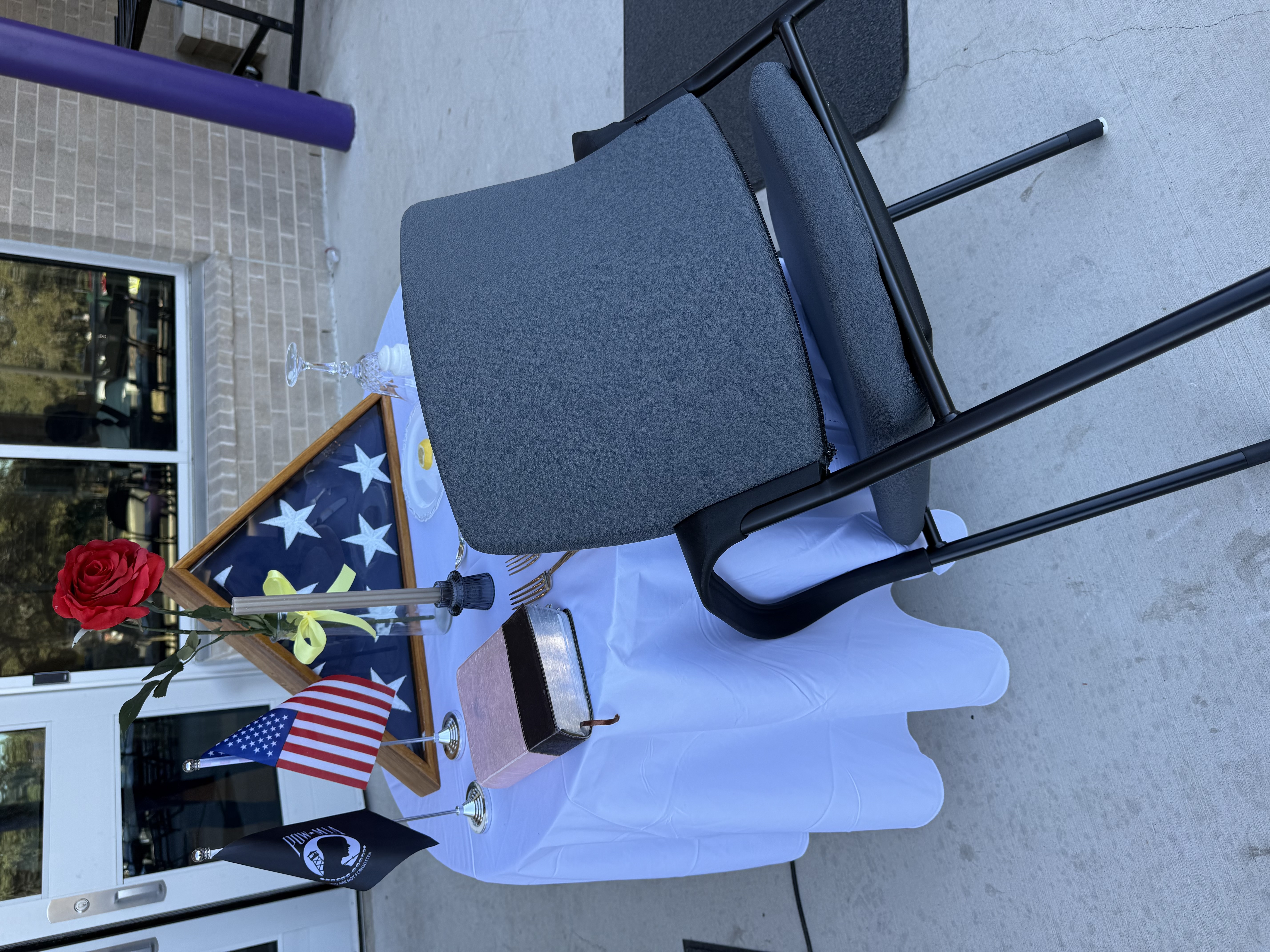 picture of american flag in case with flowers and candles on table with chair leaning agaisnt