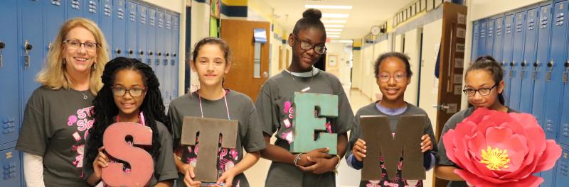 Teacher and 5 girls standing in a school hallway handing letters to spell STEM.