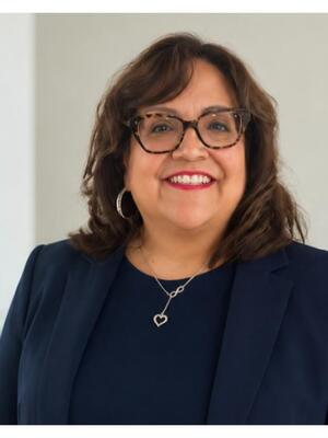 Portrait of Mrs. Benitez  with shoulder-length dark brown hair wearing tortoiseshell glasses, silver hoop earrings, a heart-shaped necklace, and a navy blazer over a dark top. She is facing the camera against a softly blurred light-colored background.